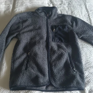 Grå fleecejacka med dragkedja - Mysig grå fleecejacka med dragkedja framtill och praktiska fickor. Perfekt för kyliga dagar. Jackan har en hög krage och en bröstficka med dragkedja för extra förvaring.