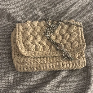 Beige väska från H&M - Säljer en snygg beige väska från H&M med flätat mönster och en kedja som axelrem. Väskan är kompakt och stilren, vilket gör den lätt att matcha med olika stilar. Lite smutsig inuti men de är inget som märks. Aldrig använd av mig men använd några få gånger av min storasyster.               Finns på Vinted oxå!!