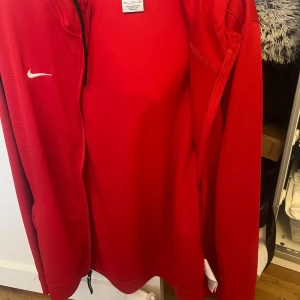 Röd jacka från Nike - Snygg röd jacka från Nike med dragkedja och långärmad design. Perfekt för en sportig look eller en avslappnad dag. Den har en liten Nike-logga på bröstet.