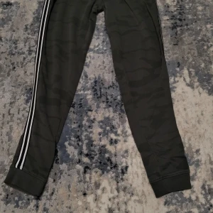 Gröna mjukisbyxor med kamouflagemönster - Snygga gröna mjukisbyxor med diskret kamouflagemönster och vita ränder längs sidorna. Perfekta för en avslappnad stil. Byxorna har ribbade muddar vid bensluten för extra komfort.