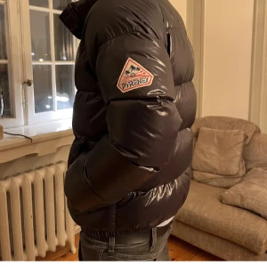 Svart pufferjacka från Pyrenex - Snygg svart pufferjacka från Pyrenex med glansig finish. Jackan har en dragkedja framtill och långa ärmar. Perfekt för kyliga dagar med sin varma och vadderade design. Märkeslogga på ärmen ger en extra touch av stil. Jag säljer nu denna jacka eftersom jag inte har användning av något vinterjacka då jag och min familj skall flytta utomlands