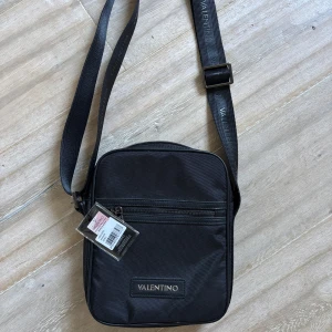 Svart axelväska från Valentino - Snygg svart axelväska från Valentino med justerbar axelrem och dragkedja framtill. Perfekt för att bära dina nödvändigheter med stil. Väskan har en elegant design med märkets logotyp på framsidan. OANVÄND