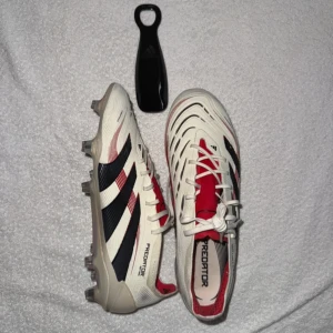 Adidas Predator fotbollsskor - Snygga Adidas Predator fotbollsskor i vitt med röda och svarta detaljer. Skorna har snörning och en aggressiv design med texten 'PREDATOR' på sulan. Perfekta för fotbollsplanen med sin stilrena look och funktionalitet.