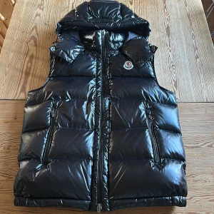 Svart dunväst från Moncler - Snygg svart dunväst från Moncler med glansig yta och dragkedja framtill. Västen har en huva och Moncler-loggan på bröstet. Perfekt för kyliga dagar när du vill hålla stilen.