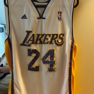 Vit Lakers basketlinne från Adidas - Säljer ett vitt Lakers basketlinne från Adidas med nummer 24 och texten 'Bryant' på ryggen. Linnet har lila och gula detaljer samt NBA-logga på framsidan. Perfekt för basketfans! Storlek M.  Lite sprickor på nummer och bokstäver annars i okej skick! 