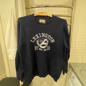 Mörkblå tröja från Lexington - Säljer en mörkblå tröja från Lexington med vit text och emblem på framsidan. Tröjan har rund hals och långa ärmar, perfekt för en avslappnad stil. Den är tillverkad i ett mjukt material som ger en bekväm passform.