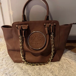 Brun handväska med kedjedetaljer - Snygg brun handväska med guldfärgade kedjedetaljer och ett elegant präglat mönster på framsidan. Väskan har två handtag och en dragkedja på baksidan. Insidan är rymlig med röd foder och flera fack för enkel organisering.