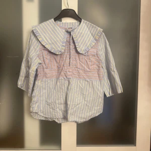 Randig blus med volangkrage - Charmig blus med randigt mönster i blått och orange. Den har en stor volangkrage och trekvartsärmar. Perfekt för en unik och stilfull look.