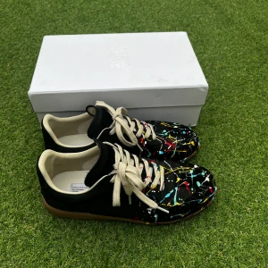 Maison Margiela Paint Splatter - Snygga sneakers från Maison Margiela med ett unikt färgstänk-mönster i flera färger på svart bas. Skorna har vita snören och en bekväm innersula. Perfekta för dig som vill sticka ut med stil. Skorna är i bra skick, om du har frågor är det bara att skriva!