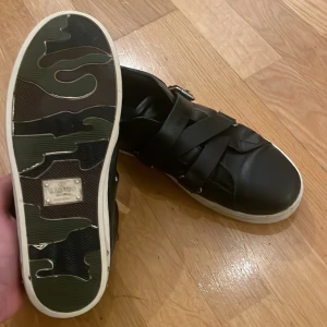 Valentino flycrew v-punk - Snygga svarta sneakers från Valentino med en unik camouflagemönstrad sula. Skorna har breda remmar över ovandelen och en diskret logotyp på insidan. Perfekta för en stilren look.