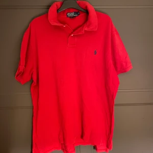 Röd pikétröja från Ralph Lauren - Säljer en klassisk röd pikétröja från Ralph Lauren med den ikoniska loggan broderad på bröstet. Tröjan har korta ärmar och en stilren krage. Perfekt för en avslappnad men stilfull look.