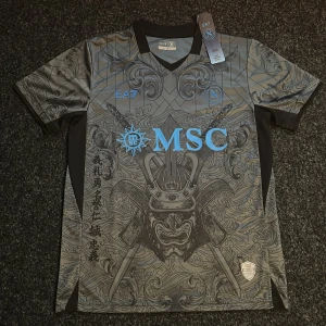 Grå och svart fotbollströja från EA7 - Snygg grå och svart fotbollströja från EA7 med detaljer i blått. Tröjan har ett unikt mönster med samurajmotiv.