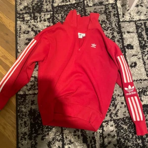 Röd tröja med dragkedja från Adidas - Säljer en snygg röd tröja från Adidas med dragkedja och klassiska vita ränder på ärmarna. Tröjan har en hög krage och Adidas-loggan på bröstet och ärmen. Perfekt för en sportig look!