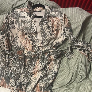 Mönstrad klänning med knytband - Snygg klänning med tropiskt mönster i gröna och beige toner. Den har långa ärmar och ett knytband i midjan för en smickrande passform. Perfekt för en stilfull look.