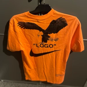 Orange t-shirt med tryck från Nike - Cool orange t-shirt från Nike med ett stort örntryck och texten 'LOGO' på baksidan. Framsidan har ett tryck som liknar en smoking med fluga. Perfekt för dig som vill sticka ut med en unik stil!