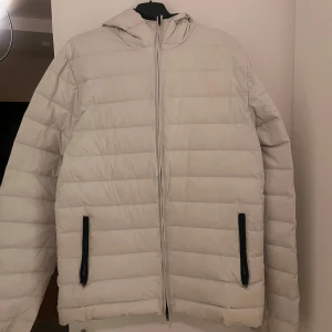 Vit dunjacka från J Lindeberg  - Säljer en stilren vit dunjacka från J.Lindeberg. Jackan har en praktisk huva och dragkedja framtill. Två fickor med svarta detaljer ger en snygg kontrast. Perfekt för kyliga dagar.