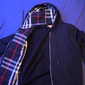 Svart hoodie från Burberry - Säljer en stilren svart hoodie från Burberry med dragkedja och klassiskt rutigt foder i huvan. Hoodien har långa ärmar och en broderad logga på bröstet. Perfekt för en casual look!