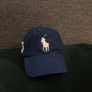 Blå keps från Ralph Lauren - Snygg blå keps från Ralph Lauren med broderad logga framtill samt 3an är broderad. Justerbar läder rem baktill för perfekt passform. Tillverkad i bomull för en bekväm känsla. perfekt nu till sommaren. Självklart är priset inte hugget Sten.