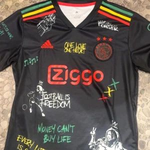 Svart fotbollströja strorlek M/L från Adidas med Bob Marley-tema - Cool svart fotbollströja från Adidas med Bob Marley-tema. Tröjan har färgglada detaljer i rött, gult och grönt samt texten 'One Love One Heart'. Baksidan pryds av ett stort porträtt av Bob Marley med texten 'Live the life you love'.
