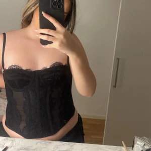 Svart spetskorsett - Snygg svart korsett med spetsdetaljer och justerbara axelband. Den har en elegant design med hakknäppning bak och genomskinliga partier som ger en sexig look. Perfekt för att lyfta din outfit till nästa nivå.
