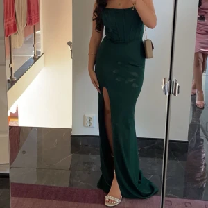 Mörkgrön långklänning - Elegant mörkgrön axelbandslös långklänning med slits framtill. Perfekt för festliga tillfällen. Klänningen har en figurnära passform och ger en sofistikerad look.