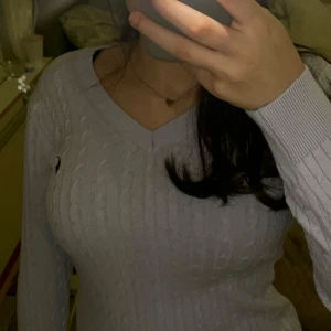 Ljuslila stickad polo tröja - Säljer en ljuslila stickad polo tröja med v-ringning och långärmad design. Tröjan har ett diskret märke på bröstet och ribbade detaljer vid ärmslut och nederkant. Perfekt för kyligare dagar!