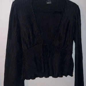 Svart ribbad blus från Gina Tricot - Snygg svart ribbad blus från Gina Tricot med v-ringning och knytning framtill. Blusen har långa ärmar och en lätt croppad design med volangdetaljer vid ärmslut och nederkant.