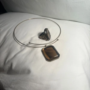 Elegant smyckesset med ring och halsband - KAN KÖPAS SEPARAT OCKSÅ! Snyggt smyckesset bestående av en ring och ett halsband. Ringen har en stor, oval mörk sten infattad i en enkel metallram. Halsbandet har en tunn metallvira med ett rektangulärt hänge i samma mörka nyans som ringen. Perfekt för att ge en annorlunda touch till din outfit.