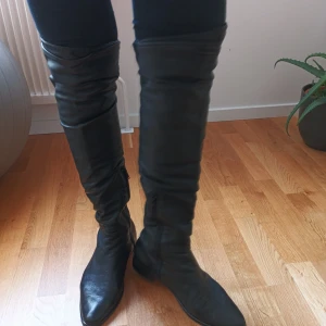 Svarta höga stövlar - Snygga svarta stövlar i skinn som går över knäna. De har en elegant och stilren design med en spetsig tå. Perfekta för att ge en trendig touch till din outfit.