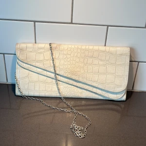 Vit kuvertväska med kedja jätte ljus beige nästan vitt  - Snygg vit kuvertväska med krokodilpräglat mönster och en elegant kedja. Väskan har en stilren design med ett blommigt foder på insidan och stängs med en tryckknapp. Perfekt för att ge en touch av elegans till din outfit.
