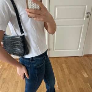 Svart axelväska med krokodilmönster - Snygg svart axelväska med krokodilmönster och justerbar rem. Perfekt för att ge en stilren touch till din outfit. Väskan har en kompakt design som gör den lätt att bära med sig.