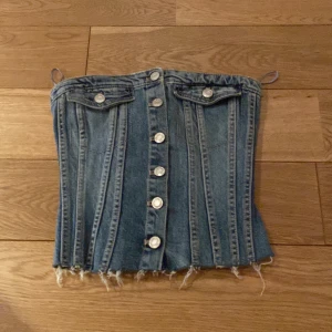 Jeanskorsett från Zara - Snygg jeanskorsett från Zara med knappar framtill och fransig nederkant. Den har dekorativa fickor och en elastisk rygg för bättre passform. Perfekt för en trendig look!
