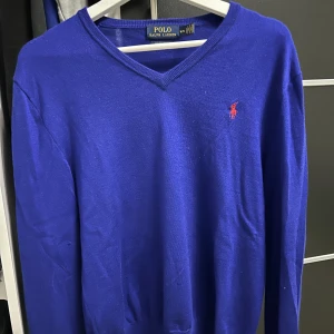 Blå tröja från Polo Ralph Lauren - Säljer en stilren blå tröja från Polo Ralph Lauren i merinoull. Tröjan har en v-ringad design och det klassiska röda logotypbroderiet på bröstet. Perfekt för en elegant och avslappnad look.