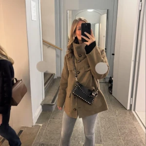 Beige ullkappa med knappar från twist and tango  - Snygg beige kappa i ull med stora knappar och hög krage. Jackan har en stilren design med två stora fickor framtill och en rak passform. Perfekt för kyliga dagar.
