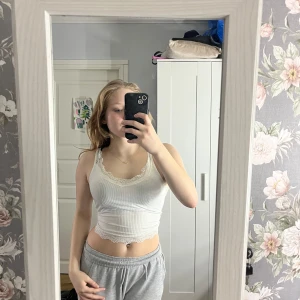 Vit ribbad linnetopp med spetsdetaljer - Säljer en söt vit ribbad linnetopp med spetsdetaljer längs kanterna. Toppen är ärmlös och har en tight passform, perfekt för sommardagar.