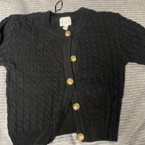 Svart kofta från H&M - Säljer en stilren svart kofta från H&M med snygga guldfärgade knappar. Koftan har ett klassiskt flätstickat mönster och ribbade kanter. Perfekt för att hålla sig varm och trendig.