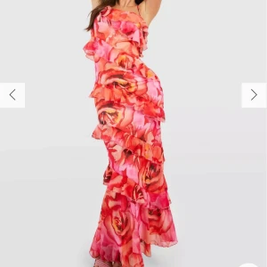 Blommig långklänning med volanger - Säljer en färgglad långklänning med ett blommigt mönster i rosa och orange toner. Klänningen har en oneshoulder-design med volanger som ger en romantisk känsla. Perfekt för festligheter! Aldrig använd