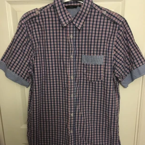 Rutig kortärmad skjorta i storlek L - Snygg rutig skjorta i blått, vitt och rött från DBC. Skjortan har korta ärmar och en bröstficka med kontrasterande mönster. Perfekt för en avslappnad stil.