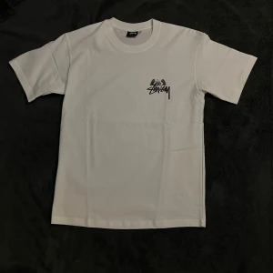 Vit t-shirt från Stüssy - Snygg vit t-shirt från Stüssy med tryck på både fram- och baksidan. Framsidan har en liten logga på bröstet medan baksidan har ett större grafiskt tryck med en ängel och logga. Perfekt för en avslappnad stil.