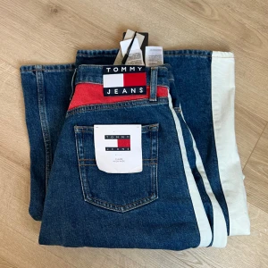 Blå jeans från Tommy Jeans - Snygga blå jeans från Tommy Jeans med vita sidoremsor och röd detalj vid midjan. De har en klassisk femficksdesign och hög midja. Supersnygga och helt oanvända!