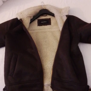 Brun jacka med teddyfoder från Identity - Säljer en snygg brun jacka från Identity med mjukt teddyfoder. Jackan har dragkedja och spännen vid kragen och nederkanten för extra stil. Perfekt för kyliga dagar!
