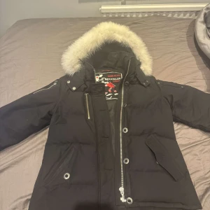 Svart dunjacka från Moose Knuckles Äkta/kvitto finns - DIGITALT KVITTO FINNS!!!Snygg svart dunjacka från Moose Knuckles med pälskantad huva och dragkedjor. Perfekt för kalla dagar med sin varma och stilrena design. Jackan har långa ärmar och en logotyp på ärmen.