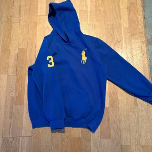 Blå hoodie från Ralph Lauren - Snygg blå hoodie från Ralph Lauren med gult polospelarbroderi på bröstet och siffran 3 på ärmen. Perfekt för en avslappnad stil med en klassisk touch. Den har en praktisk magficka och en bekväm huva.