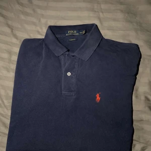 Mörkblå pikétröja från Ralph Lauren - Säljer en klassisk mörkblå pikétröja från Ralph Lauren med röd broderad logotyp på bröstet. Tröjan har korta ärmar och en traditionell krage med knappar. Perfekt för en stilren och avslappnad look ☺️.