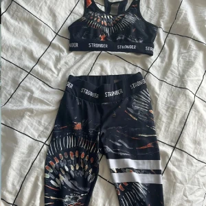 Svart träningsset från Stronger  - Snyggt svart träningsset från Stronger med ett unikt mönster i orange, vitt och grått. Setet består av en sport-bh och matchande tights, båda med elastisk midja och logotypdetaljer. Perfekt för träning och att känna sig stark och bekväm.