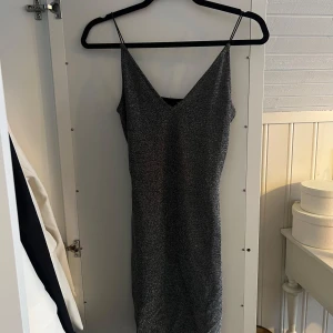 Glittrig klänning med smala axelband - Snygg glittrig klänning i silver med smala axelband och v-ringning. Perfekt för festliga tillfällen. Klänningen har en figurnära passform och är i ett glittrande material som fångar ljuset fint.