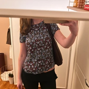 Mönstrad blus med korta ärmar - Säljer en blus med ett detaljerat mönster i rött, blått och vitt. Blusen har korta ärmar och en rund halsringning. Toppen har knappdetaljer och är i storlek Xs/S. Väldigt bra skick. Materialet är viskos