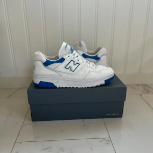 Vita och blå sneakers från New Balance - Snygga vita sneakers med blå detaljer från New Balance. Skorna har en klassisk design med snörning och märkets logga på sidan. Perfekta för en stilren look. I princip helt nya köptes på footlocker i italien.