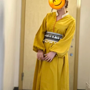 Gul långklänning med dekorativt bälte - Elegant gul långklänning med v-ringning och långa ärmar. Klänningen har ett dekorativt bälte med mönster och detaljer i svart och vitt, vilket ger en unik touch. Perfekt för speciella tillfällen.