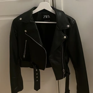 Svart skinnjacka från Zara - Snygg svart skinnjacka från Zara med dragkedjor och bälte. Jackan har en klassisk bikerstil med silverfärgade detaljer och är perfekt för en cool look💕